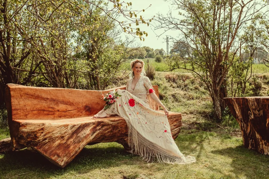 A bride in a boho fringed gown with floral embroidery, sitting outdoors in a Poppy Perspective dress