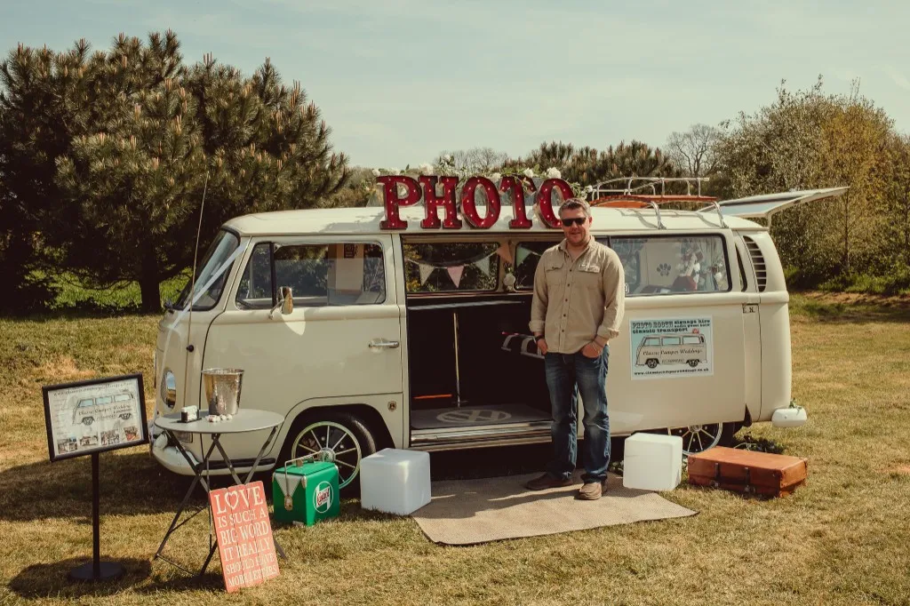 A cream VW camper van photo booth with PHOTO marquee letters, set up outdoors for a wedding