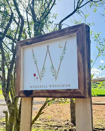 The wooden Woodhill Weddings entrance sign with a floral W monogram among the trees at the venue in Faversham, Kent